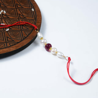 A handcrafted Rubellite Freshwater Pearl Rakhi displayed on a wooden platform with decorative elements in the background.
